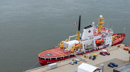 An overhead view of a bright red and white Canadian Coast Guard ship featuring a helipad on its bow, securely docked at a pier next to parked vehicles and maritime equipment.