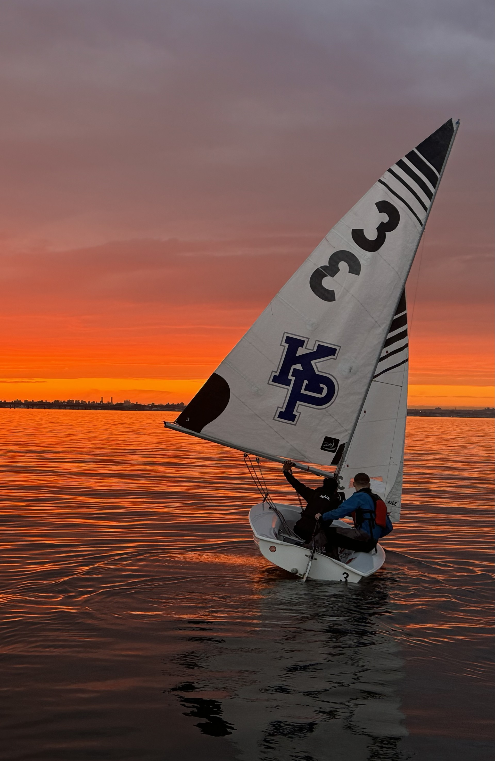 A sail boat with with a white and blue sail reading 3 3 KP on the water at sunset.