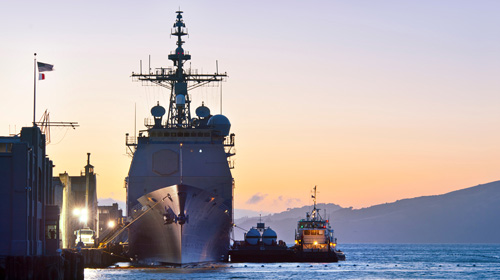 A tall, grey naval warship docked at a concrete pier at sunset, accompanied by a smaller tugboat and service barges moored directly alongside its hull.