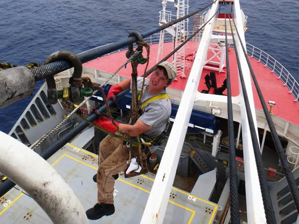Midshipman Nathanael Kutz, wearing a safety harness, soiled work clothes, and heavy red gloves, hangs suspended while working on thick steel cables high above the red deck of the MV Ocean Giant, with the open ocean visible in the background.