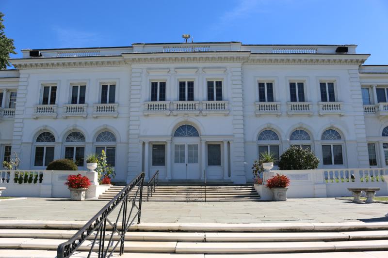 Wiley Hall at USMMA is a grand, classical-style white building featuring arched ground-floor windows and a roofline balustrade. Wide stone steps lead up past potted red flowers to the building's central double-door entrance under a clear blue sky.