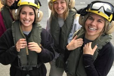 Four smiling female midshipmen posing together on a ship's deck during their Sea Year, wearing protective flight gear including yellow safety helmets with ear protection, goggles, and green inflatable life vests.