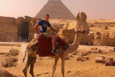 Midshipman Thomas Duncan, wearing a blue USMMA t-shirt, smiles while sitting atop a camel in the Egyptian desert. The iconic Great Sphinx and a massive Great Pyramid of Giza stand prominently in the sandy background.